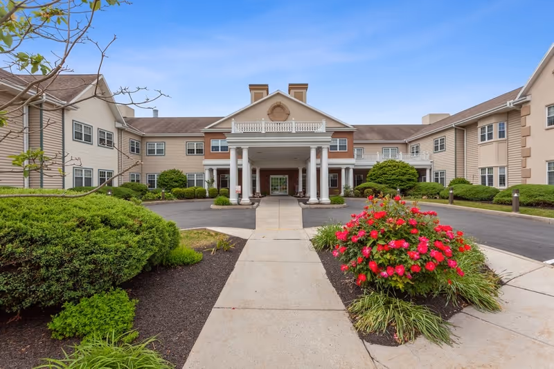 Front exterior view of The Bridges at Bent Creek facility showing a large entrance with white columns, a covered driveway, landscaped bushes, and vibrant red flowers under a clear blue sky.