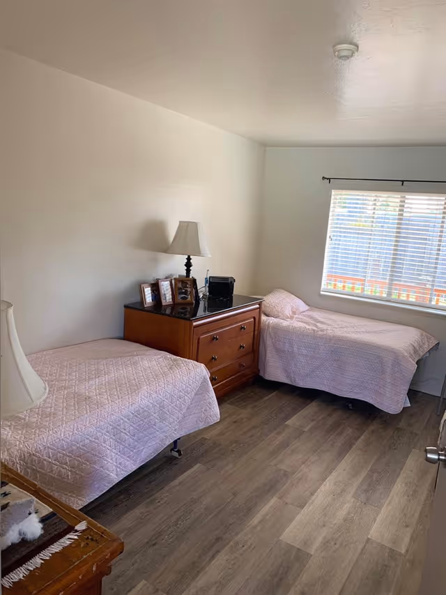 Small bedroom with two twin beds covered in pink quilts, a wooden dresser topped with a lamp and framed photos, a window with blinds, and wood-look flooring.