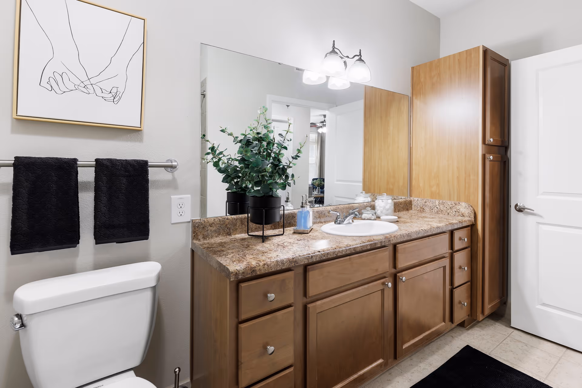 A clean bathroom with a white toilet, wooden cabinets with a granite countertop, a white sink, a large mirror, and a light fixture with three bulbs above the mirror. Two black towels hang on a towel rack next to the toilet, and a framed line drawing of two hands holding pinky fingers is on the wall. A potted green plant, soap dispenser, and cotton balls are on the countertop.