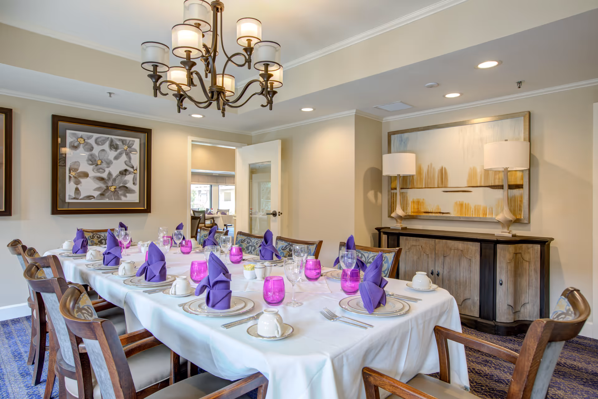 A dining room with a long table set for a meal, featuring white tablecloth, purple folded napkins, clear wine glasses, and purple water glasses. The room has wooden chairs with patterned cushions, a chandelier overhead, framed floral artwork on the wall, and a sideboard with two lamps and an abstract painting above it.