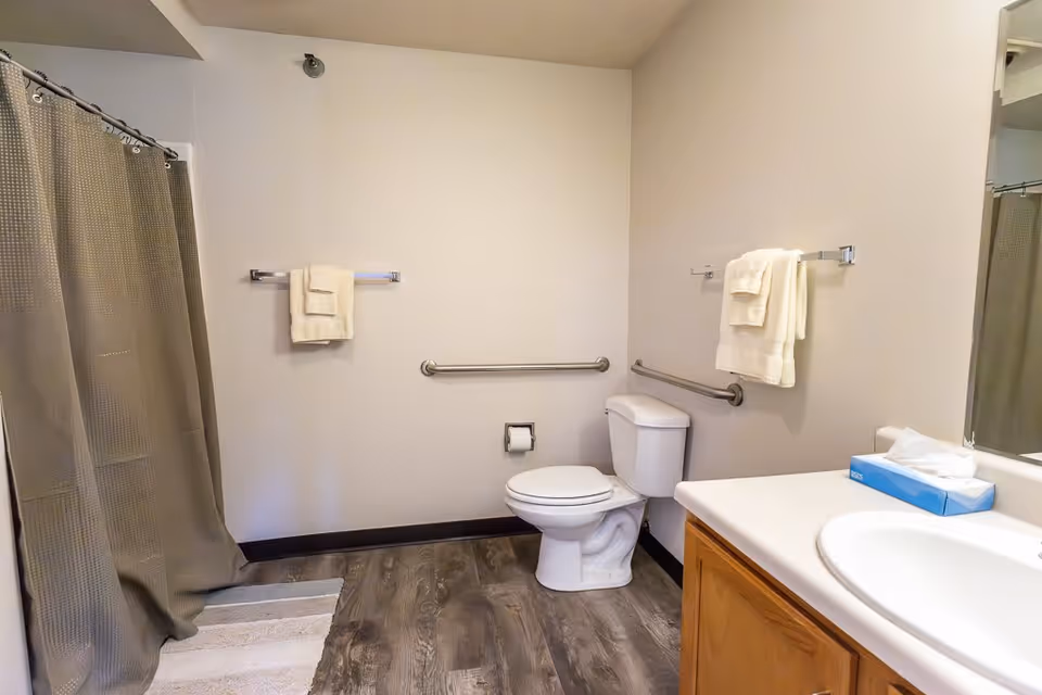A clean bathroom with a white toilet, a sink with a wooden cabinet, a mirror, a box of tissues on the counter, beige walls, a shower with a beige curtain, two towel racks with folded white towels, and a wooden floor with a white bath mat.