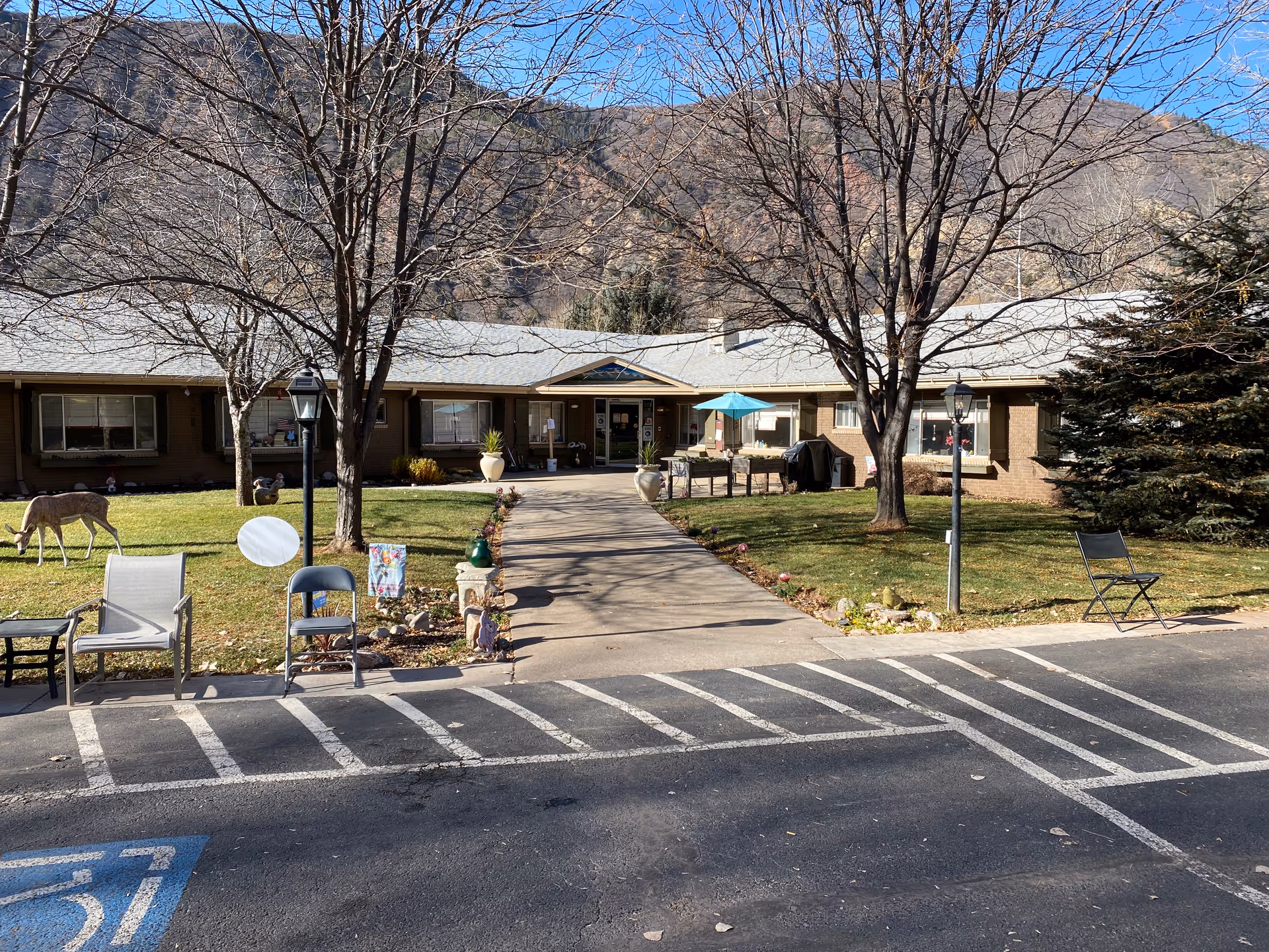 Single-story building front entrance with a paved walkway, lawn and chairs, lamp posts and bare trees, with mountains in the background.