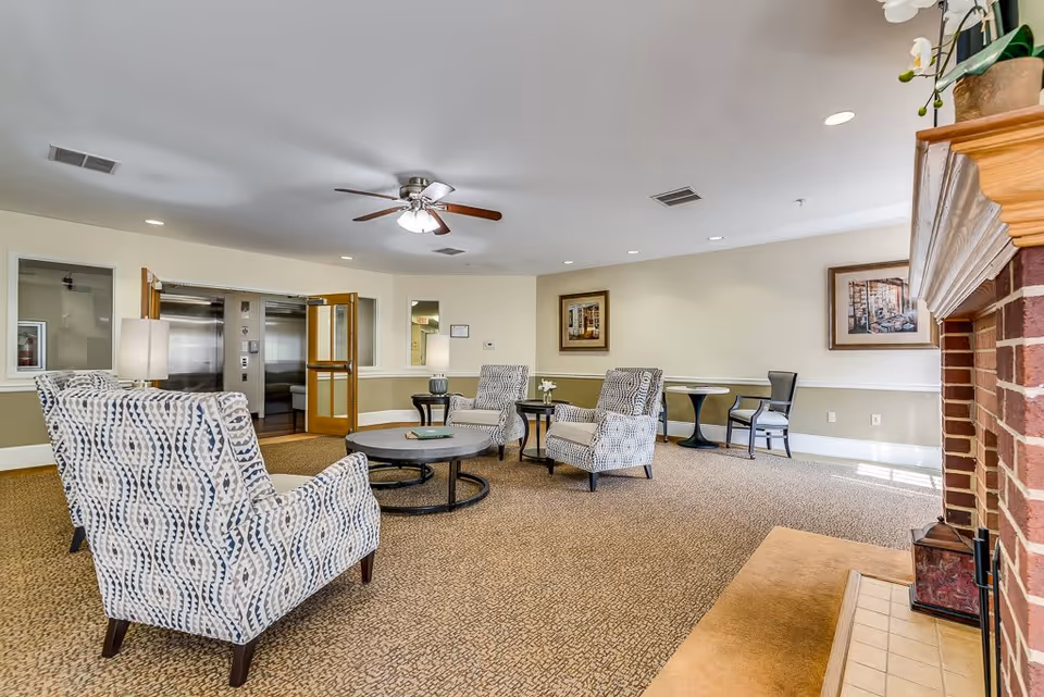 A spacious living room area in a senior living facility featuring patterned armchairs arranged around a round coffee table, a ceiling fan with lights, framed artwork on the walls, a brick fireplace on the right, and an elevator with open doors in the background.