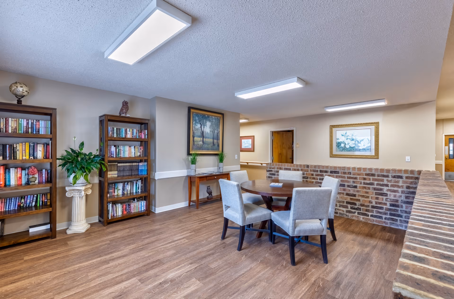 A cozy interior common area with wooden flooring, two tall bookshelves filled with books, a round wooden table surrounded by four upholstered chairs, a small wooden console table with decorative plants, and framed artwork on the walls. The space is well-lit with ceiling lights and has a partial brick wall.