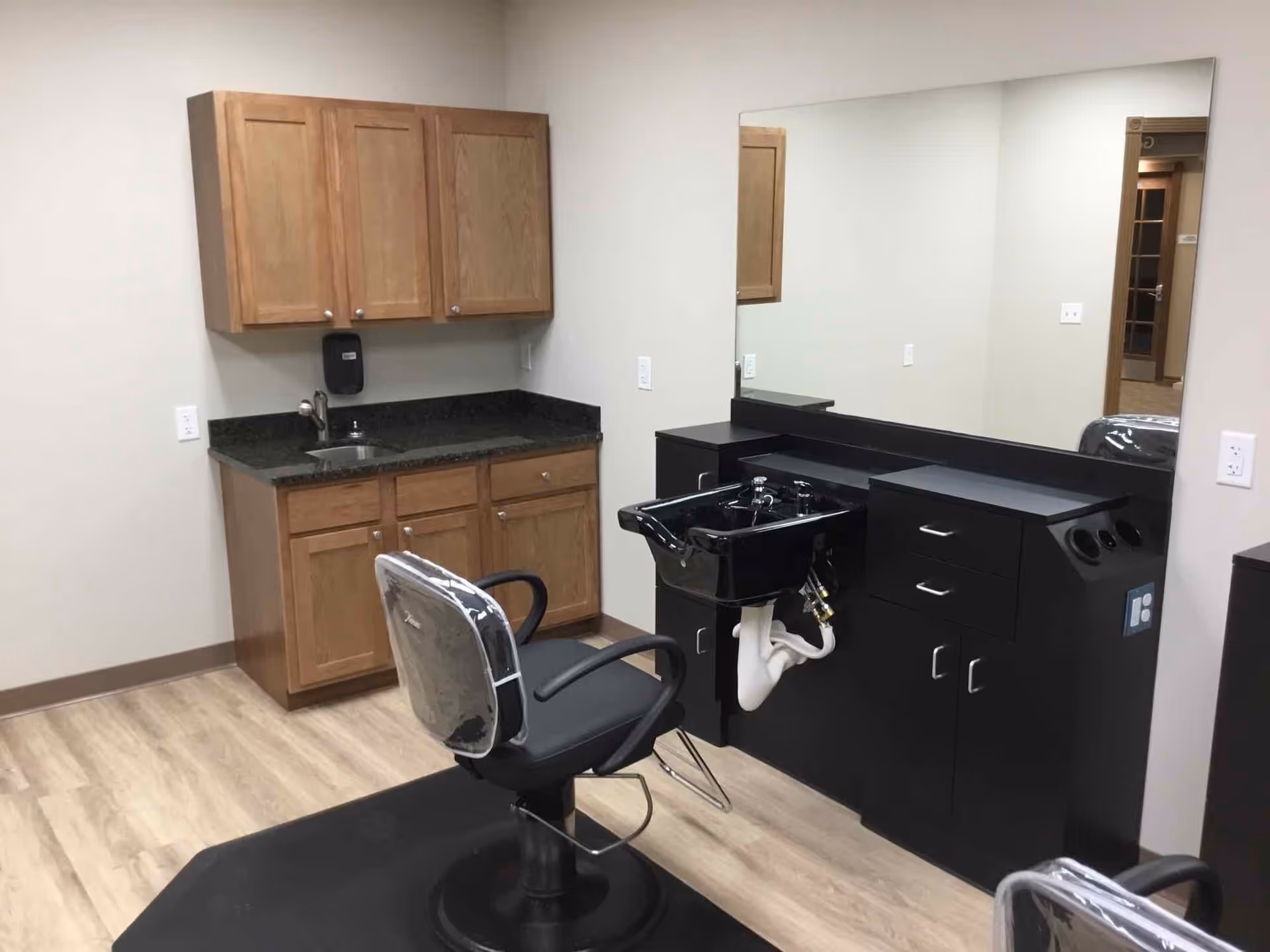 Interior of a salon area in a senior living facility featuring a black salon chair with a protective cover, a black hair washing sink with cabinetry underneath, a large mirror on the wall, and wooden cabinets with a countertop and sink in the background.