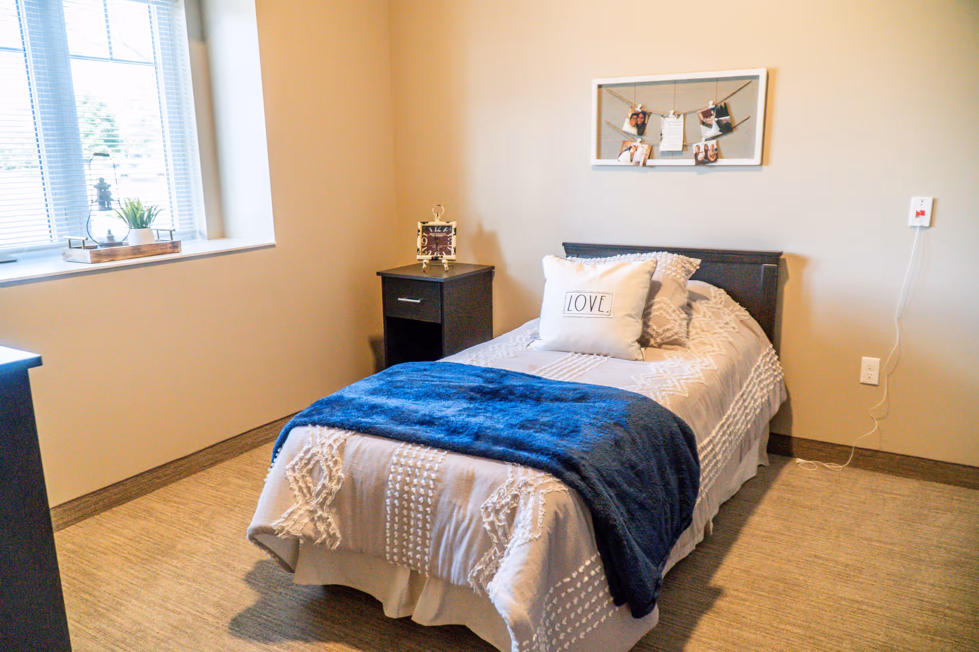A cozy bedroom with a single bed covered in a white textured bedspread and a blue throw blanket. There are two pillows on the bed, one with the word 'LOVE' printed on it. Next to the bed is a black nightstand with a decorative clock. A window with blinds lets in natural light, and a photo frame with clipped pictures hangs on the beige wall above the bed.