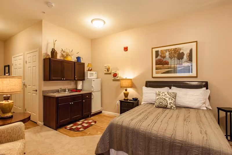 Cozy studio bedroom with a bed and nightstand beside a small kitchenette featuring dark cabinets, a sink and a mini-fridge.