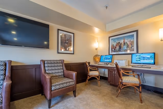 A cozy common area with two patterned armchairs and two wooden chairs with cushions placed in front of a long desk with two desktop computers and a printer. The walls are painted beige with dark wood paneling on the lower half. A flat-screen TV is mounted on the wall above one armchair, and two framed artworks hang on the walls. The room is softly lit by wall sconces.