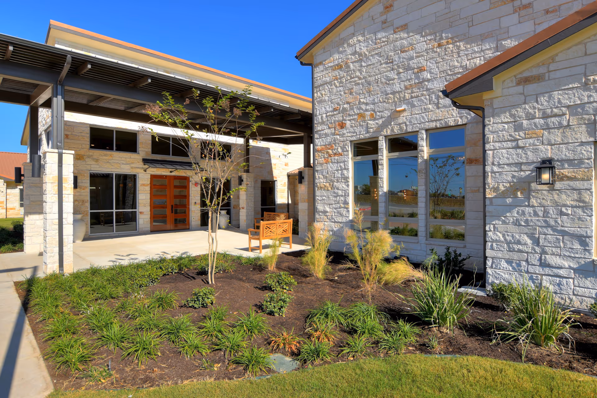 Stone-clad building entrance with a covered porch, landscaped planting bed, and a wooden bench in front of double doors.