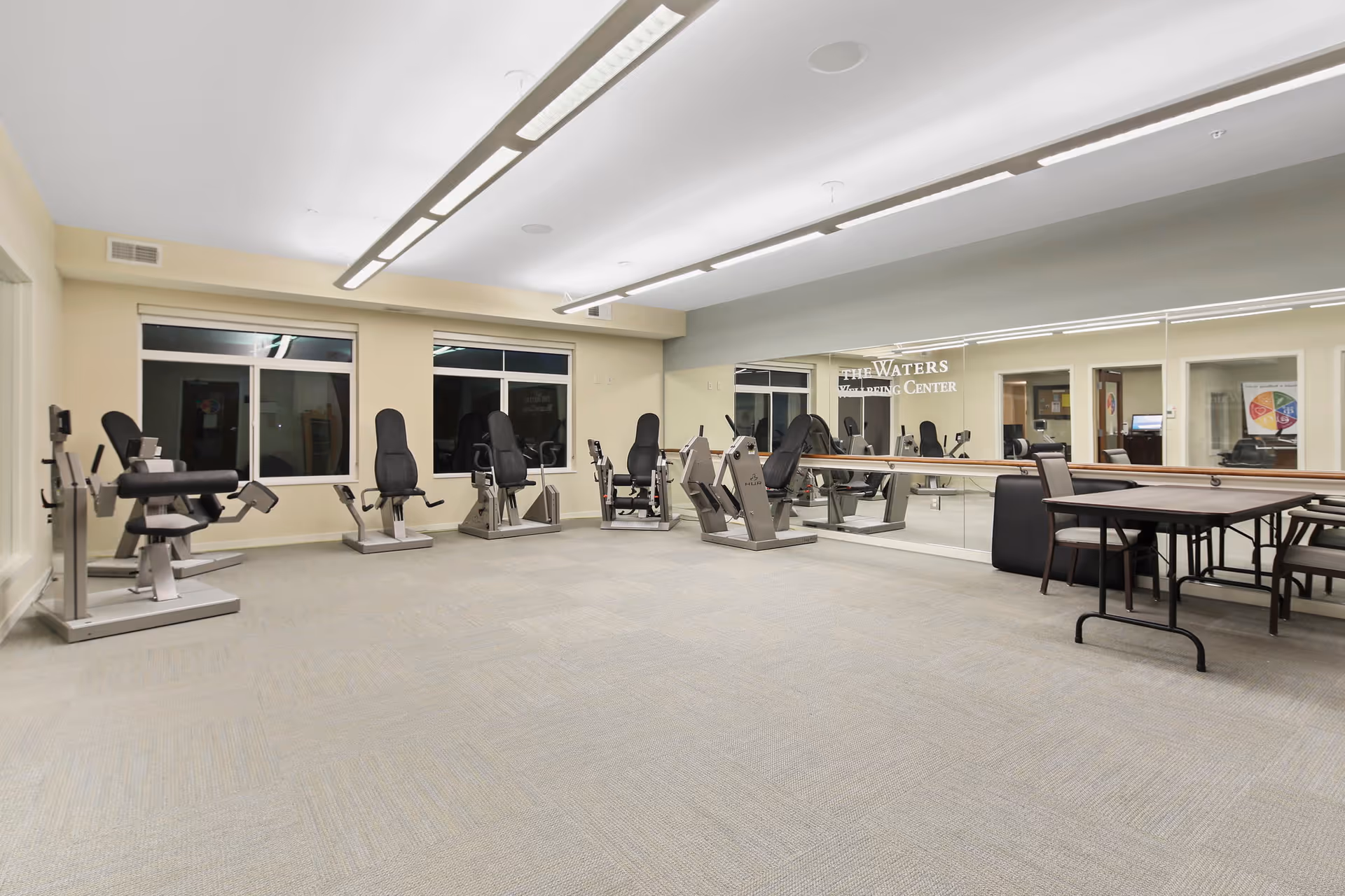A spacious exercise room with several stationary fitness machines arranged along the walls. The room has large windows, bright overhead lighting, and a wall-length mirror reflecting the equipment. There is also a table with chairs in one corner. The mirror has the text 'The Waters Wellness Center' on it.