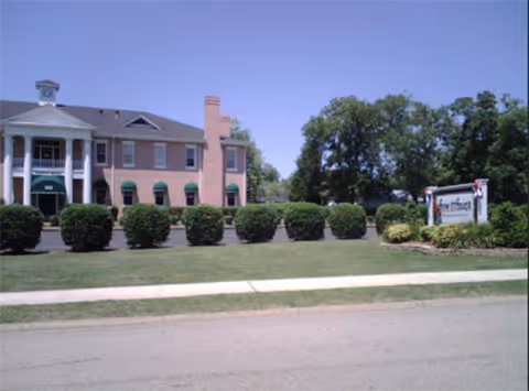 Exterior view of a two-story brick building with white columns and green awnings, surrounded by neatly trimmed bushes and a lawn. A sign near the right side of the image reads 'Keestone at Hewitt House'. Trees are visible in the background under a clear blue sky.