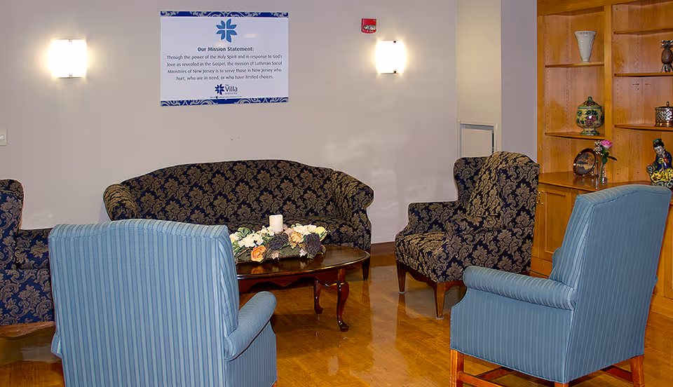 A cozy sitting area with two blue striped armchairs, two dark patterned armchairs, and a matching sofa arranged around a wooden coffee table with floral decorations and a candle. The walls are light-colored with two wall sconces providing soft lighting. A wooden shelving unit with decorative items is visible on the right side. A framed mission statement from The Villa At Florham Park is mounted on the wall.