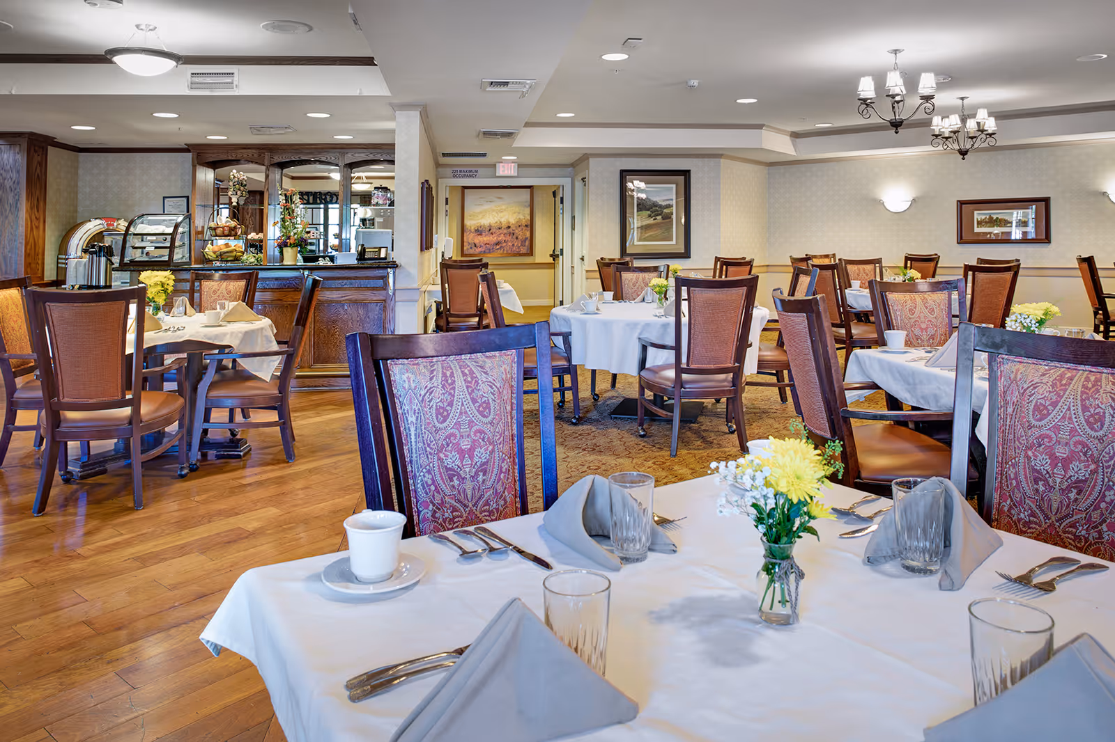 Bright dining room with round and rectangular tables set with white linens, wooden chairs, floral centerpieces, and a service counter in the background.
