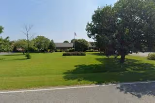 A large green lawn with several trees and shrubs in front of a single-story building with a flagpole displaying an American flag.