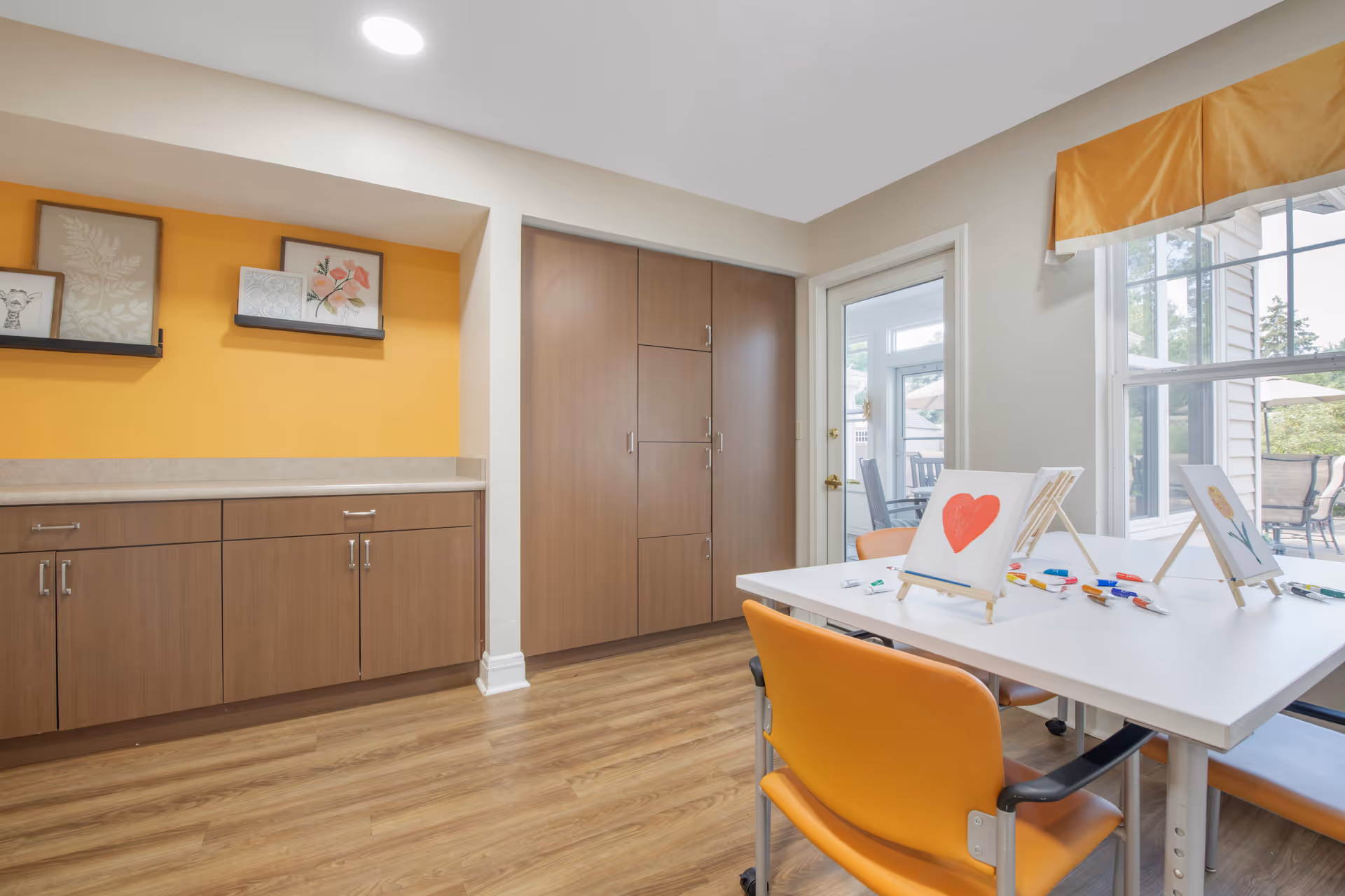 A bright room with wooden flooring and light-colored walls, featuring a white table with orange chairs around it. On the table are small easels holding paintings, including one with a red heart and another with a flower. The room has built-in wooden cabinets and shelves with framed artwork on a yellow accent wall. A glass door and large windows provide a view of an outdoor patio area with chairs and umbrellas.