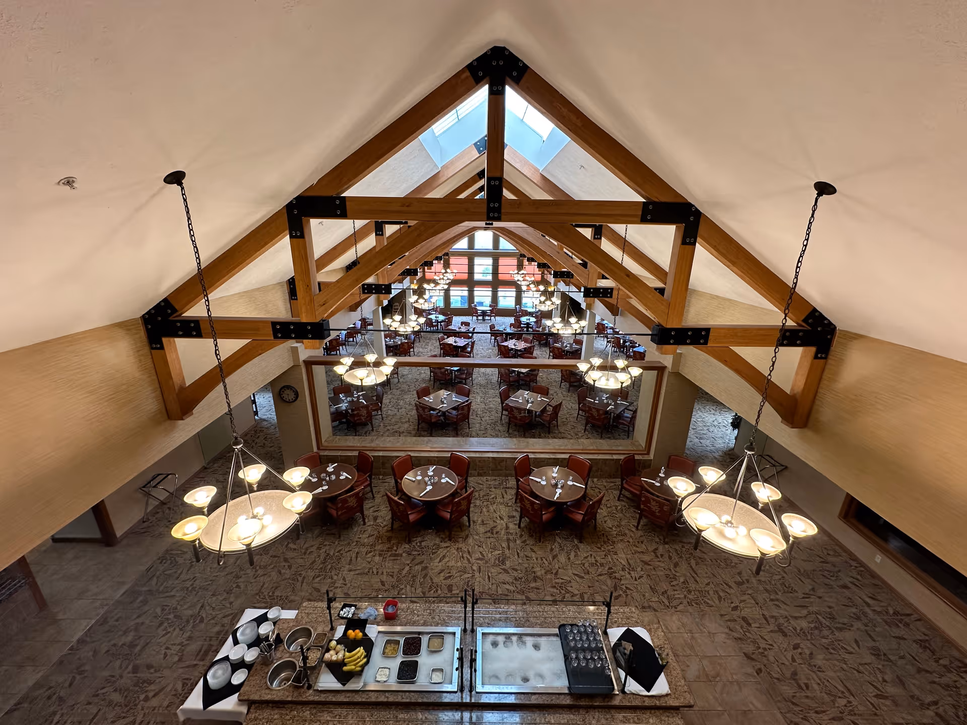 Interior view of a spacious dining room with high vaulted ceilings supported by wooden beams. Multiple round tables with chairs are arranged neatly, and a buffet station with plates, bowls, and food items is visible in the foreground. Large windows at the far end allow natural light to fill the room.