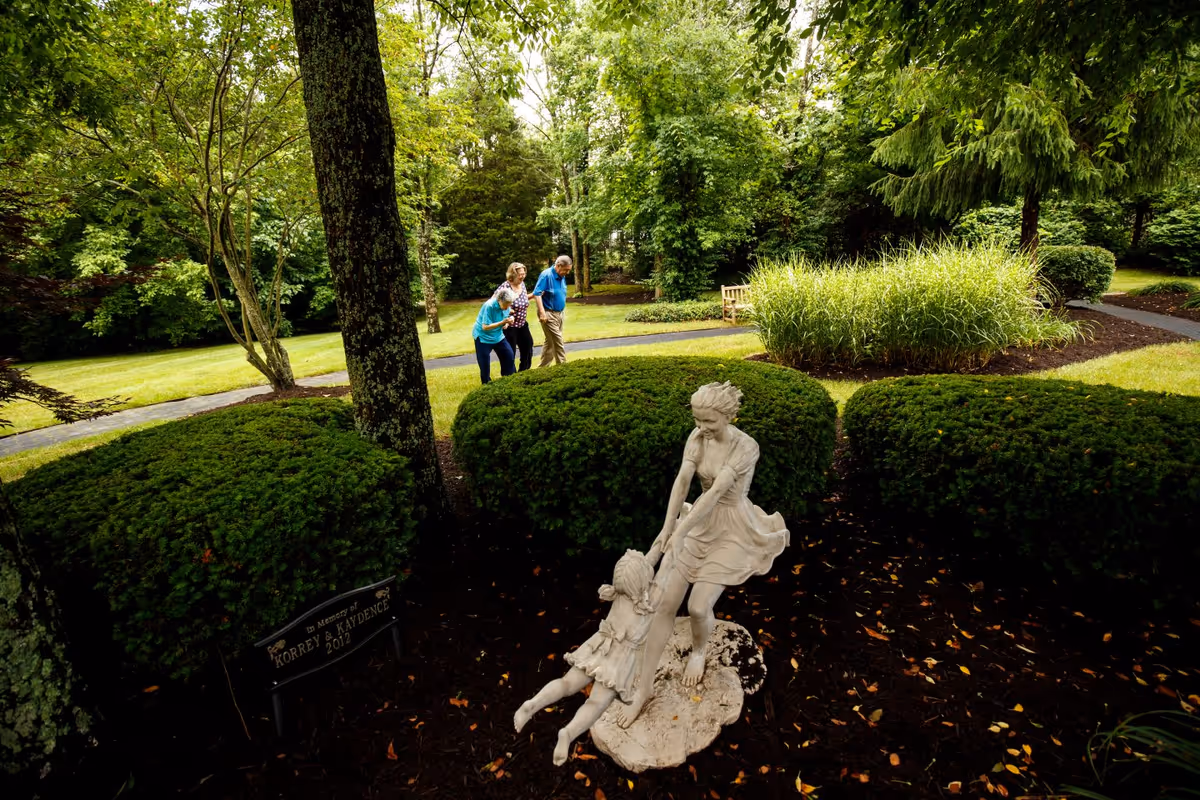 A serene outdoor garden area with lush green trees, bushes, and a winding paved pathway. In the foreground, there is a white statue of two children playing, surrounded by neatly trimmed bushes. In the background, three elderly people are walking along the path, enjoying the natural surroundings.