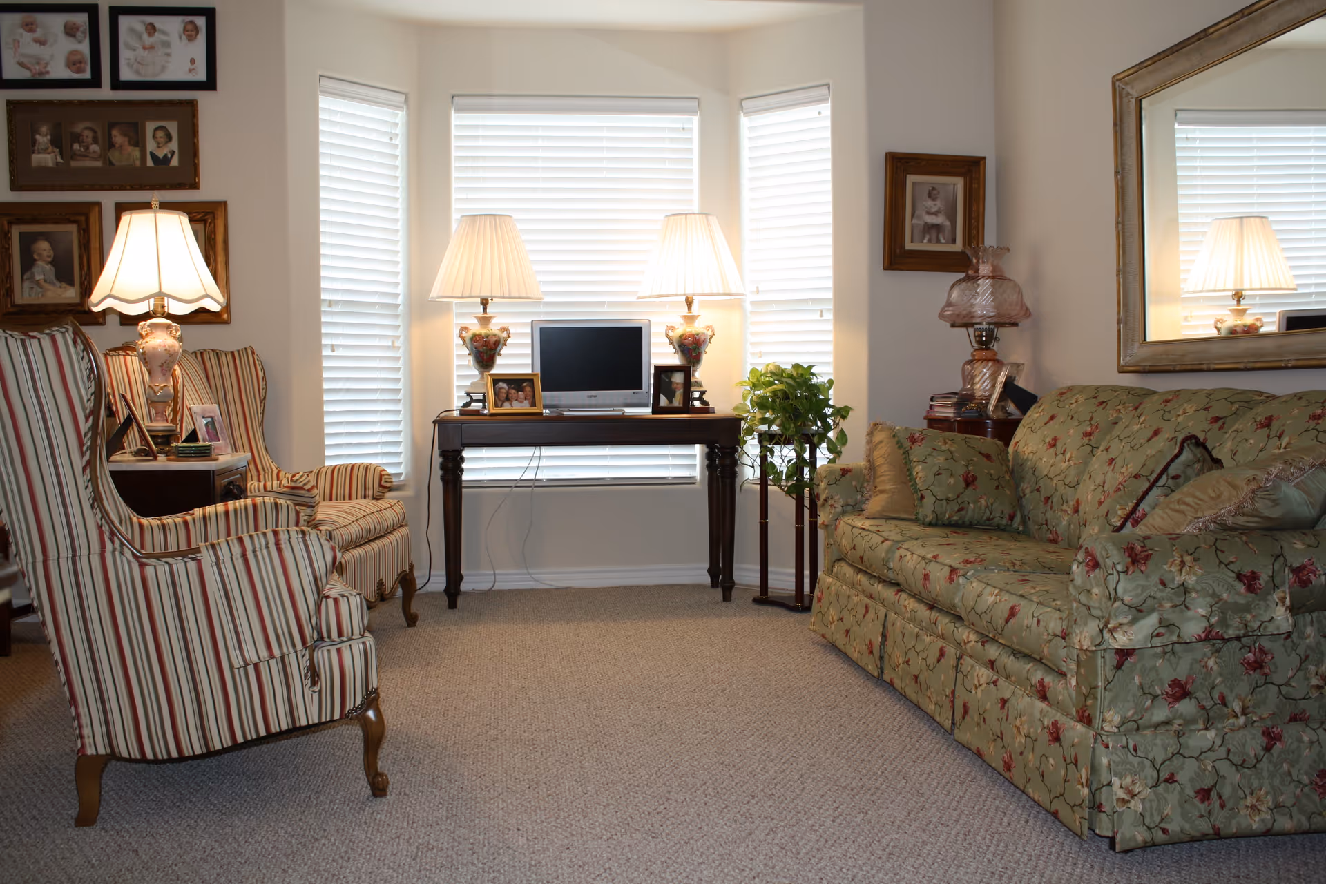 A cozy living room with a floral patterned green sofa on the right and two striped armchairs on the left. A wooden table with a small TV and two decorative lamps is positioned in front of three windows with white blinds. Family photos and framed portraits hang on the walls, and a large mirror reflects part of the room. A small plant stands next to the table.