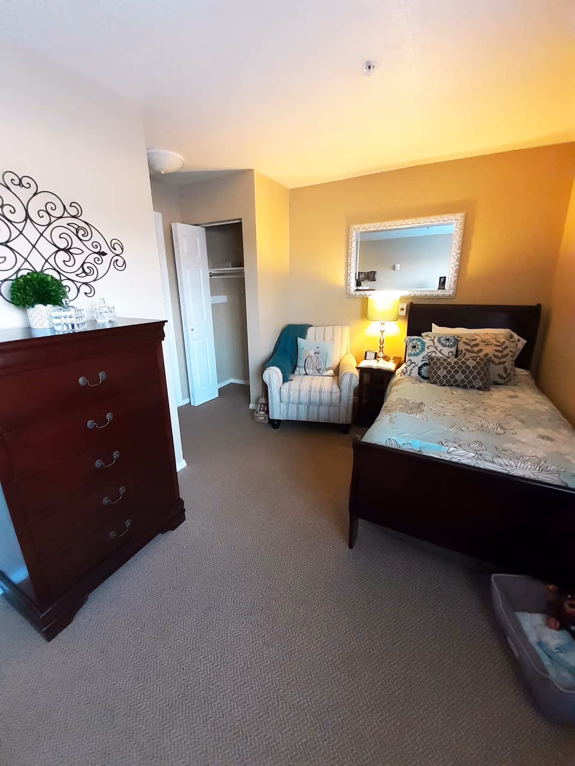 A cozy bedroom in a senior living facility featuring a single bed with patterned bedding and multiple pillows, a wooden dresser with decorative items on top, a comfortable armchair with a throw blanket and a pillow, a nightstand with a lamp, and a mirror on the wall. The room has beige walls and carpeted flooring.