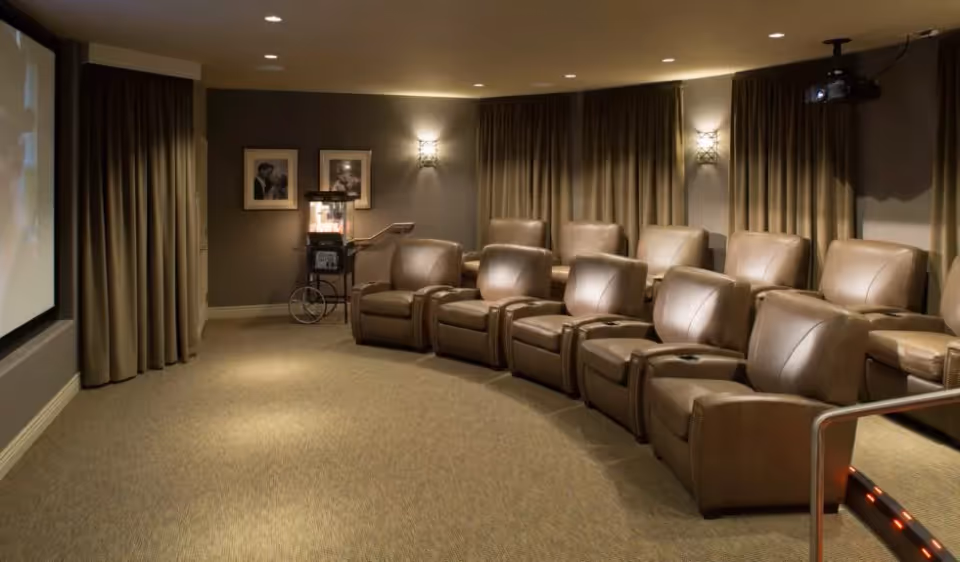 A cozy home theater room with two rows of brown leather recliner chairs facing a large projection screen. The room has beige carpeting, dark curtains covering the windows, wall sconces providing soft lighting, and a vintage-style popcorn machine in the corner.