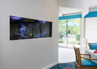 Interior view of a senior living facility showing a built-in wall aquarium with fish, a seating area with wooden chairs and teal cushions, and a glass door leading to an outdoor patio with greenery and an umbrella.