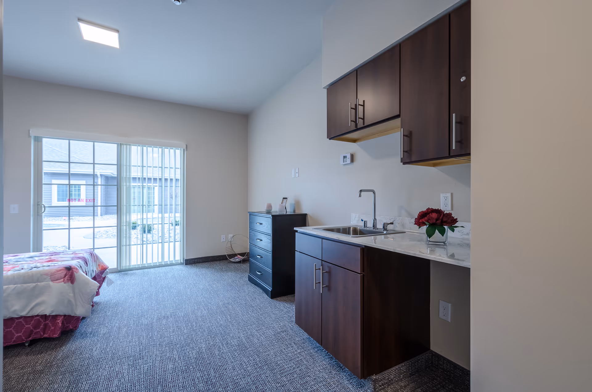 Interior view of a senior living facility room with a small kitchenette featuring dark wood cabinets and a sink on the right. A black dresser is against the far wall, and a bed with floral bedding is partially visible on the left near a sliding glass door with vertical blinds that opens to an outdoor area.