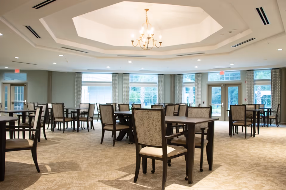 A spacious dining room with multiple dark wood tables and cushioned chairs arranged neatly on a beige carpet. Large windows with patterned curtains allow natural light to fill the room. A chandelier hangs from a recessed ceiling in the center of the room.