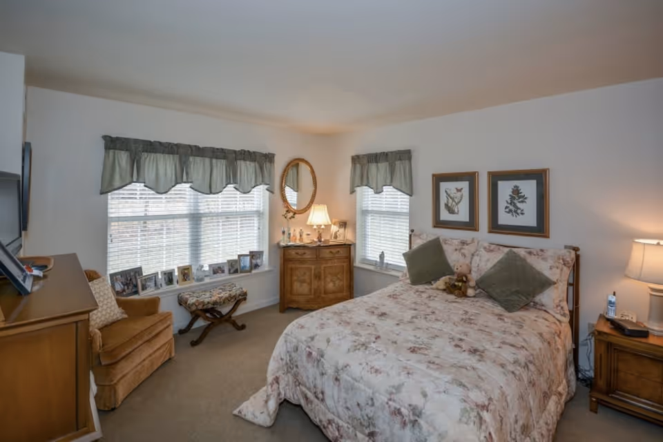 A cozy bedroom with a floral-patterned bedspread on a double bed, two green pillows, and a teddy bear. The room has two windows with green valances, a wooden dresser with a lamp and a round mirror above it, a wooden nightstand with a lamp and a phone, a cushioned armchair, and a small bench by the window. Several framed pictures are displayed on the windowsill and walls.
