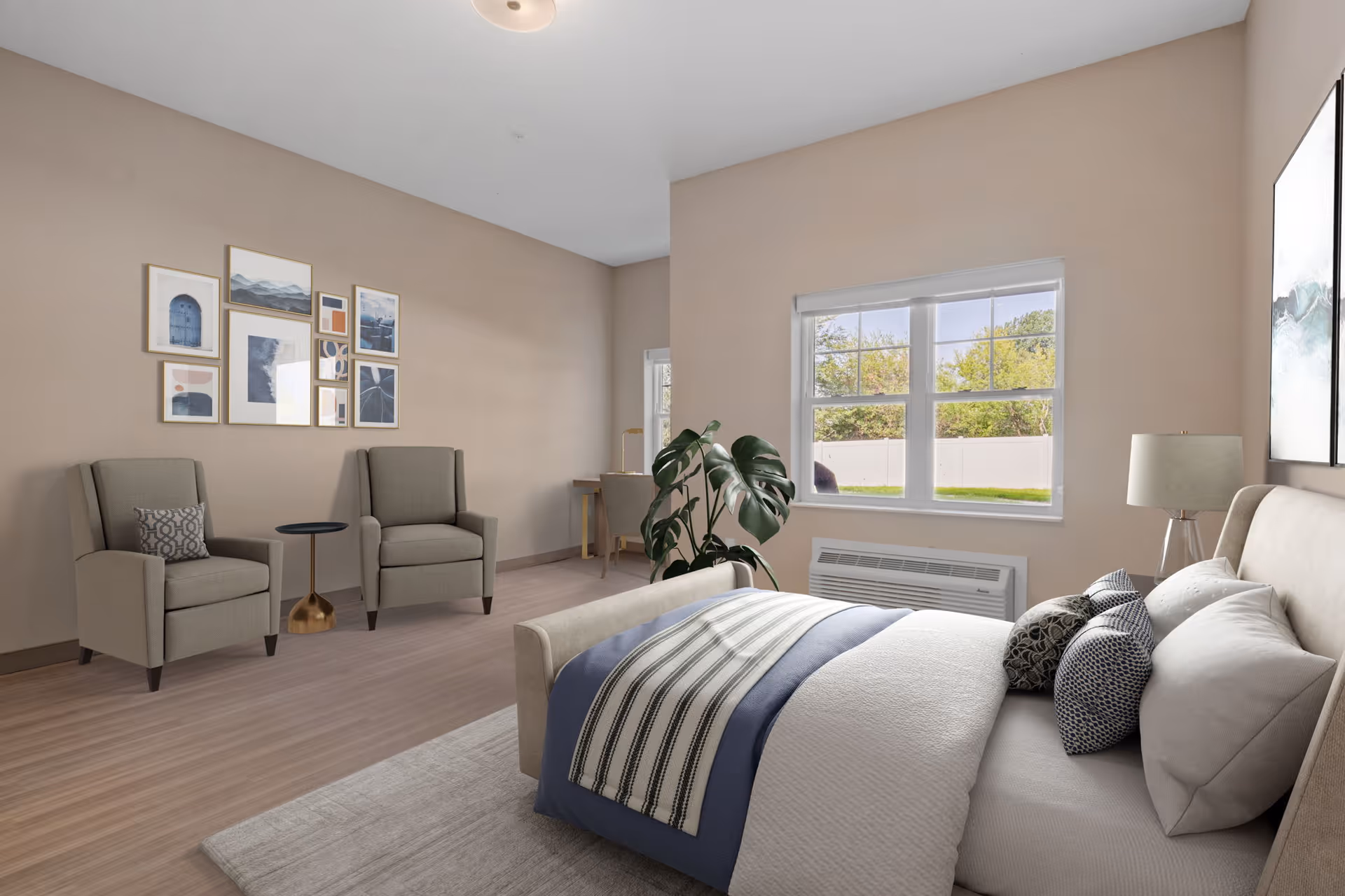 A bright bedroom with a large bed featuring multiple pillows and a striped blanket. Two armchairs with a small round table between them are positioned against a wall decorated with a gallery of framed artwork. A window shows a view of greenery outside. There is a small desk and chair in the corner, a floor lamp, and a potted plant near the window.