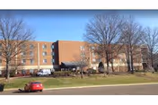 Exterior view of a multi-story brick building with several windows, a main entrance with a covered drop-off area, leafless trees in front, and a red car parked on the street nearby under a clear blue sky.