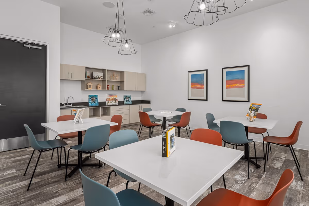 A modern dining room with several square white tables surrounded by blue and red chairs. The room has light wood flooring, white walls, and two colorful abstract paintings on one wall. There are light wood cabinets and shelves with decorative items and artwork along the back wall, and three pendant lights with wireframe shades hang from the ceiling.