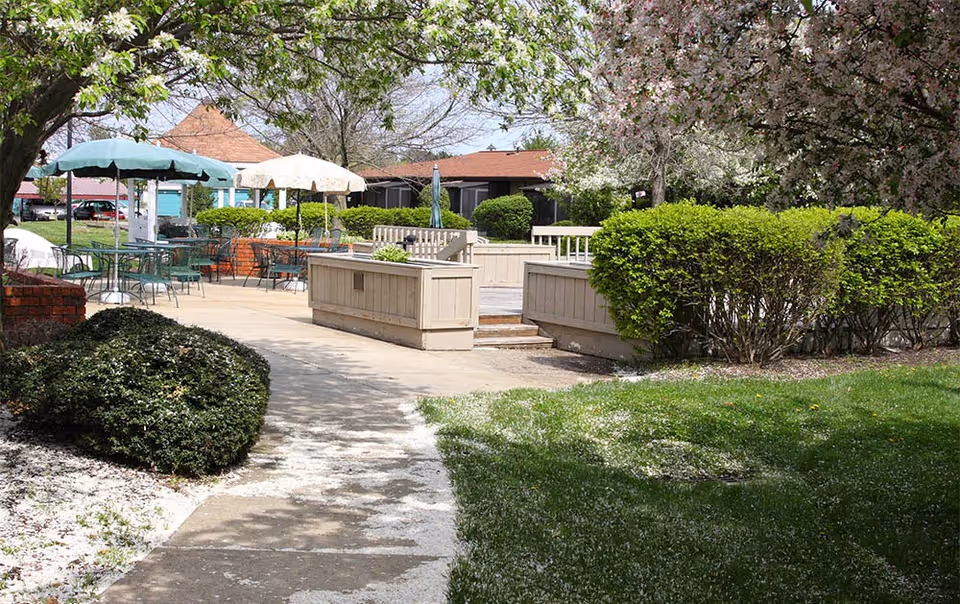 Outdoor patio area with tables and chairs under umbrellas surrounded by green bushes and blooming trees, with a paved walkway leading through the garden space.