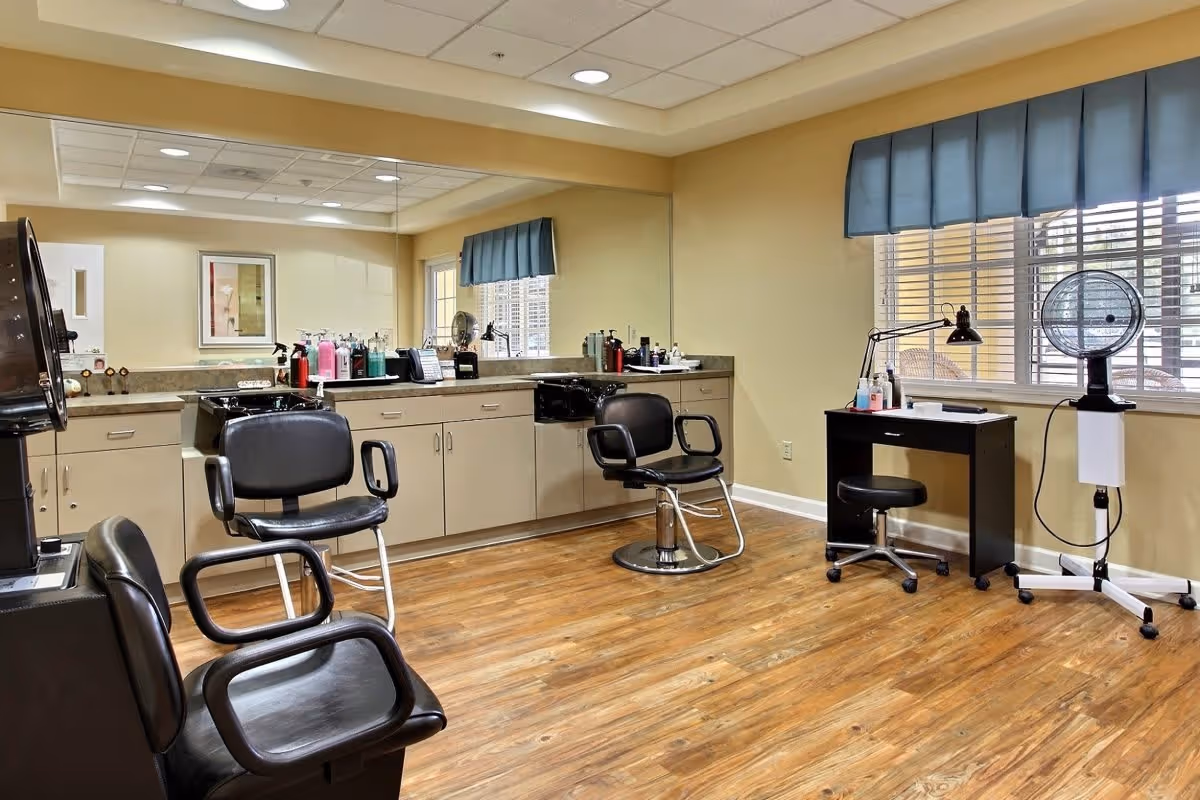 Interior view of a salon area in an assisted living community featuring three black salon chairs, a large mirror above a countertop with various hair care products, a small desk with a lamp and stool near a window with blue valance, and a hair dryer on a stand. The room has wooden flooring and beige walls.