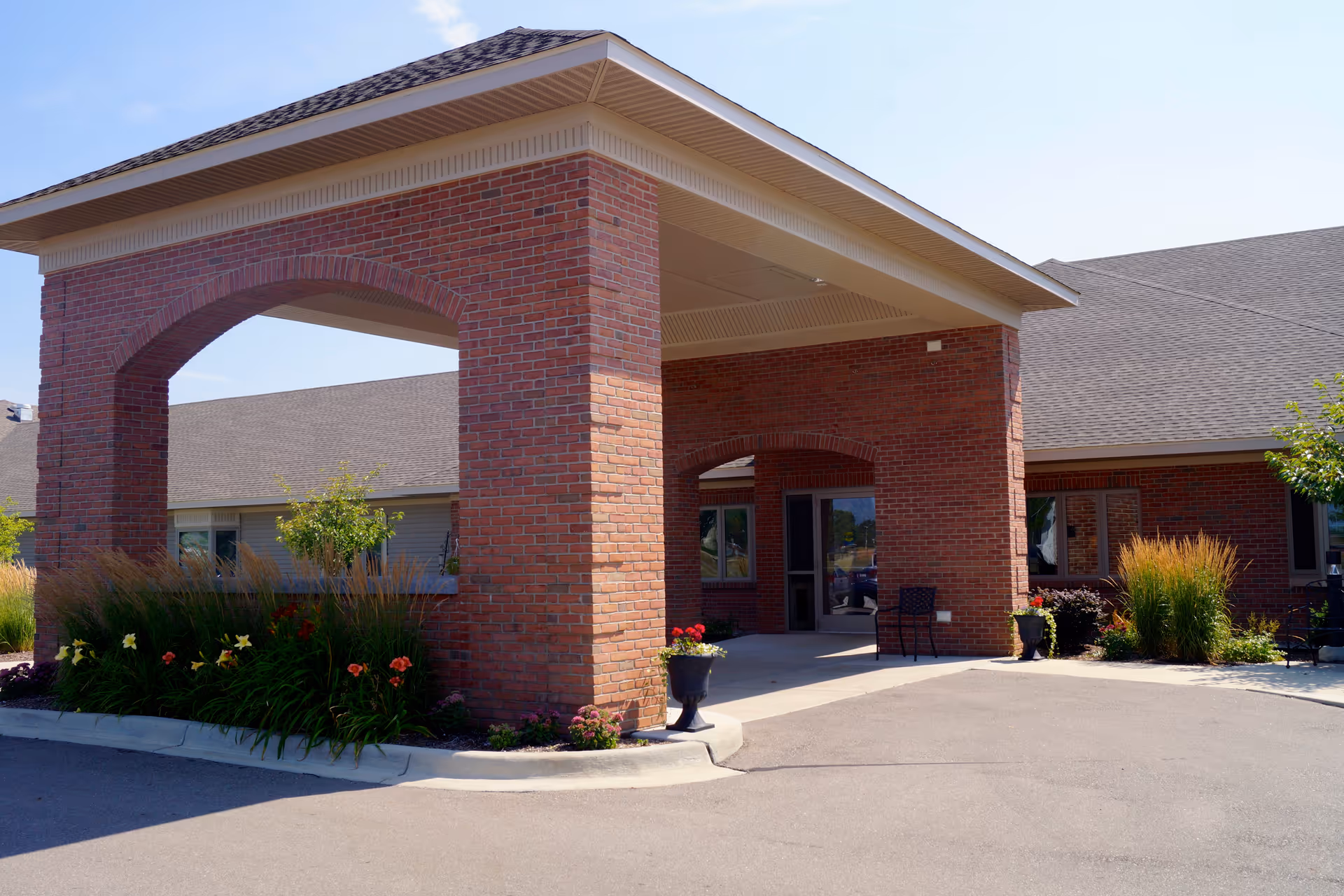 Entrance of a senior care facility with a covered drop-off area supported by brick pillars, surrounded by landscaped plants and flowers under a clear blue sky.