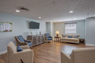 A cozy living room area with light blue walls and wood flooring. The room features a beige sofa with blue and beige cushions, two beige armchairs, two blue armchairs, a wooden side table with a lamp, a decorative cabinet with a flat-screen TV mounted above it, and a window with a valance letting in natural light.