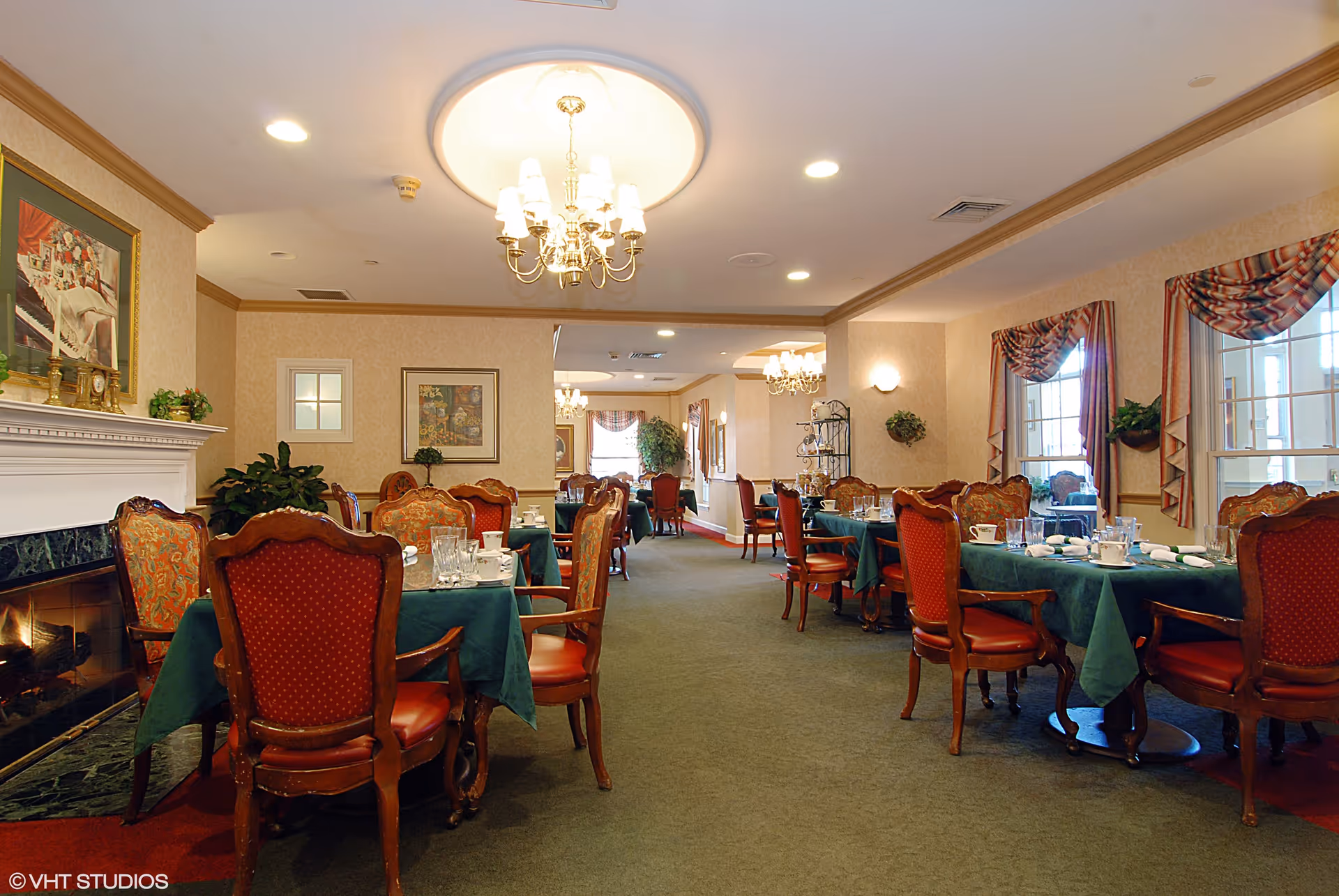 Formal dining room with tables set with green tablecloths, red upholstered chairs, chandeliers and a fireplace.