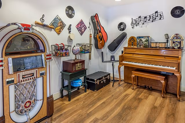 A music-themed room with a vintage jukebox on the left, a wooden upright piano on the right, and various musical decorations on the walls including vinyl records, a guitar, a banjo, and musical notes. The room has wooden flooring and a small table with a globe and other decorative items.