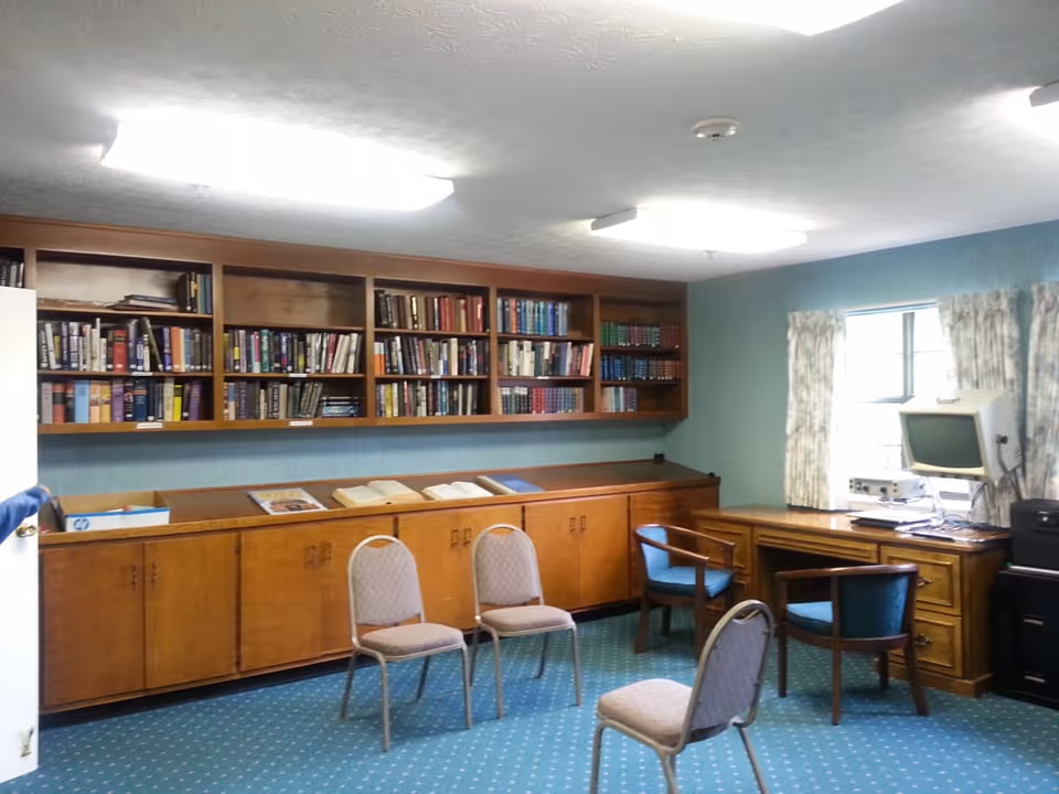 A small library or reading room with wooden bookshelves filled with books, a long wooden cabinet below the shelves, three beige chairs arranged in a semi-circle, two blue cushioned wooden chairs near a wooden desk with a computer monitor, and a window with floral curtains letting in natural light.