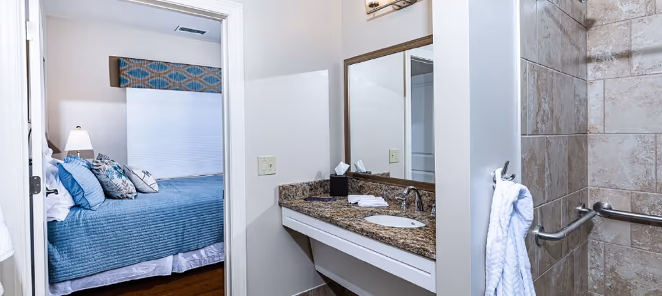 Bathroom vanity with granite countertop and mirror beside a tiled shower, with a bed visible through an open doorway.