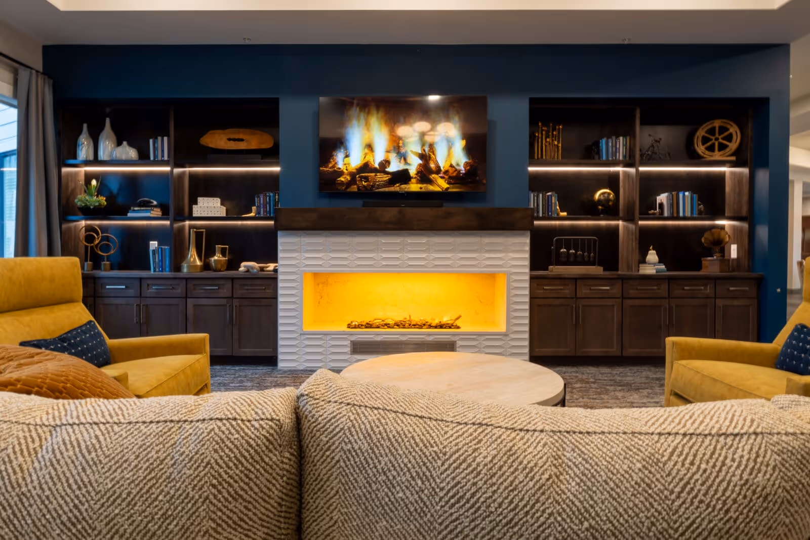 Modern lounge area with a built-in fireplace and TV flanked by illuminated shelving, and upholstered seating in the foreground.