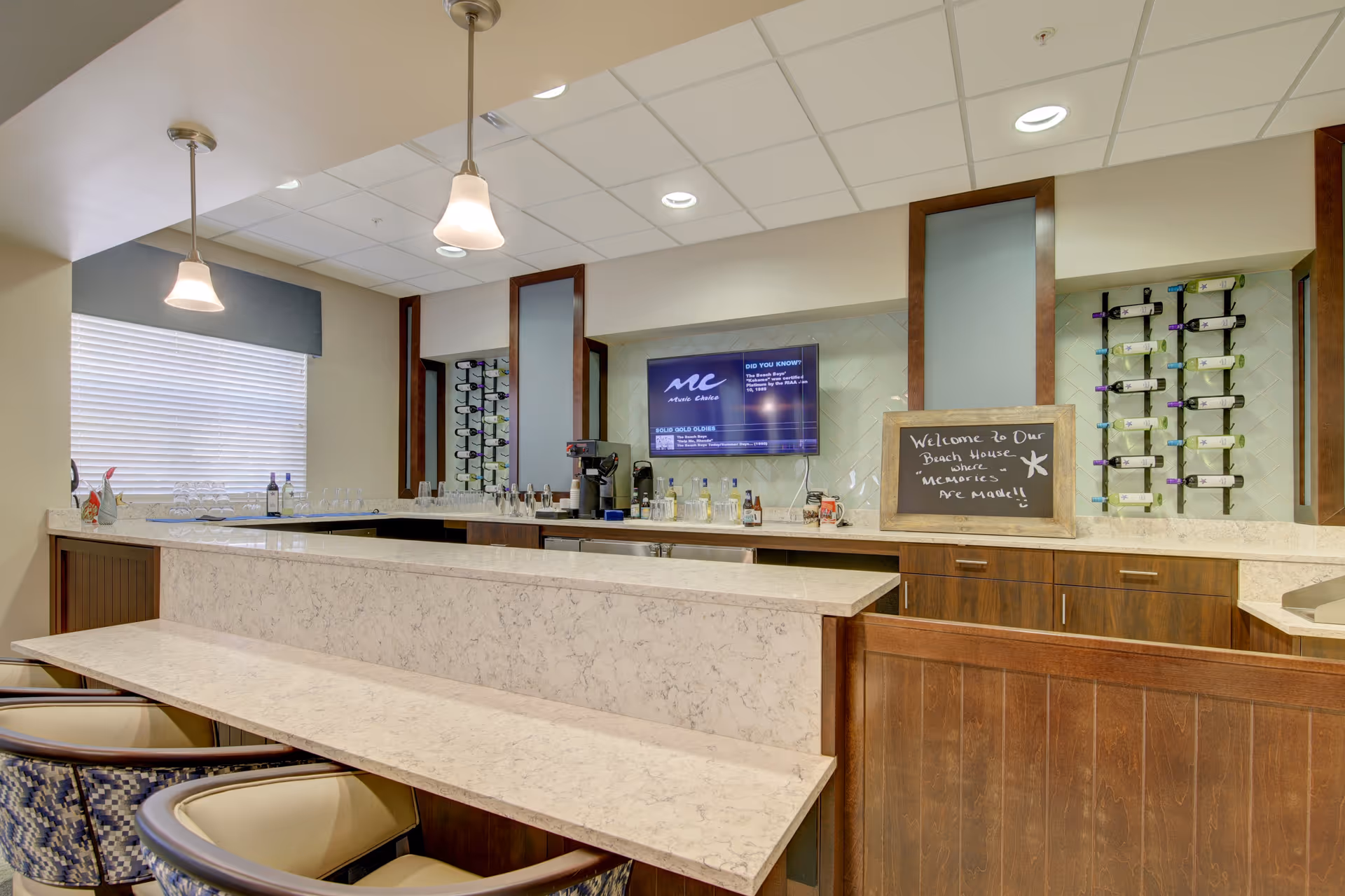 Interior view of a modern bar area in Beach House Assisted Living & Memory Care with marble countertops, wooden cabinetry, wine racks on the wall, a chalkboard sign welcoming guests, pendant lights hanging from the ceiling, and several chairs lined up along the counter.