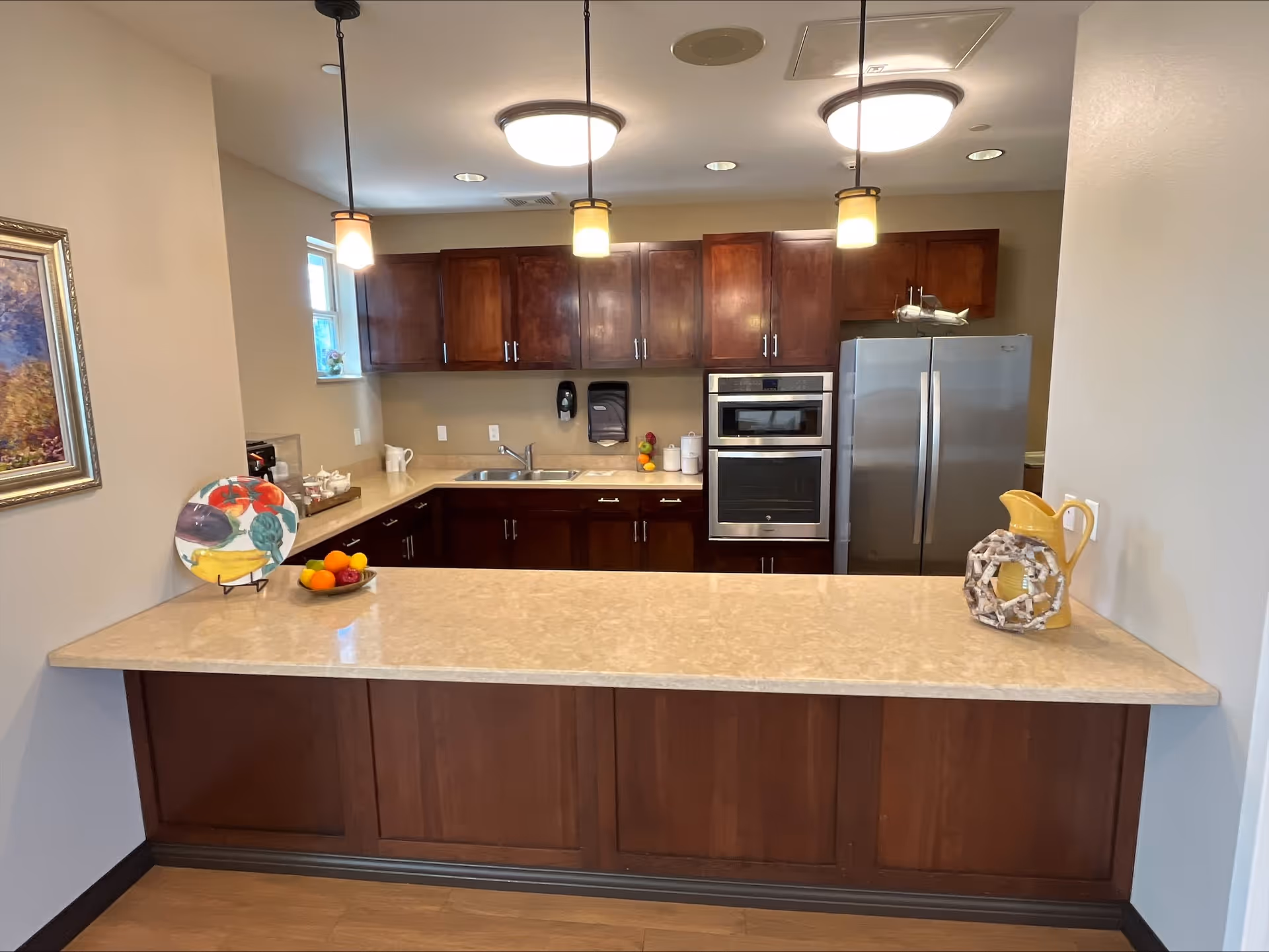 A modern kitchen with dark wooden cabinets, a beige countertop island, stainless steel refrigerator and oven, three hanging pendant lights, a small window, and decorative items including a colorful plate and a bowl of fruit.
