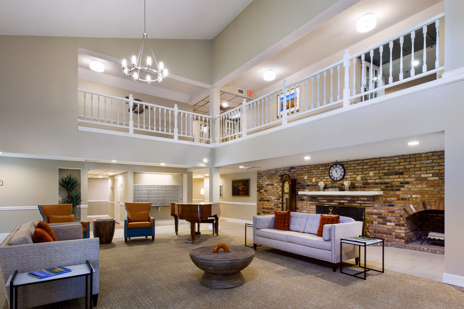 A spacious living room area in a senior living facility with two gray sofas, two orange and blue armchairs, a round wooden coffee table, and a grand piano. The room features a brick fireplace with a clock above it, a grandfather clock, and a balcony with white railings overlooking the space. The walls are painted beige, and the room is well-lit with ceiling lights and a chandelier.
