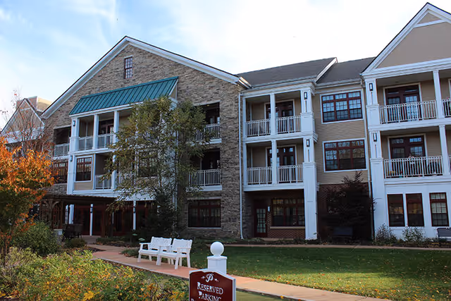 Exterior view of a multi-story senior living facility building with balconies, large windows, and a stone facade. There is a green lawn, trees, and a walkway with white benches in front. A sign indicating reserved parking is visible near the walkway.