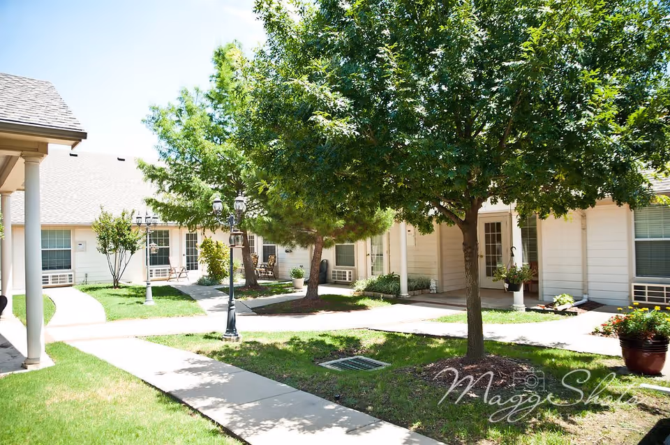 Outdoor courtyard area of Bethesda Gardens Frisco Assisted Living with green grass, trees, paved walkways, and white buildings with windows and doors surrounding the courtyard. There are some chairs and potted plants near the building entrances.