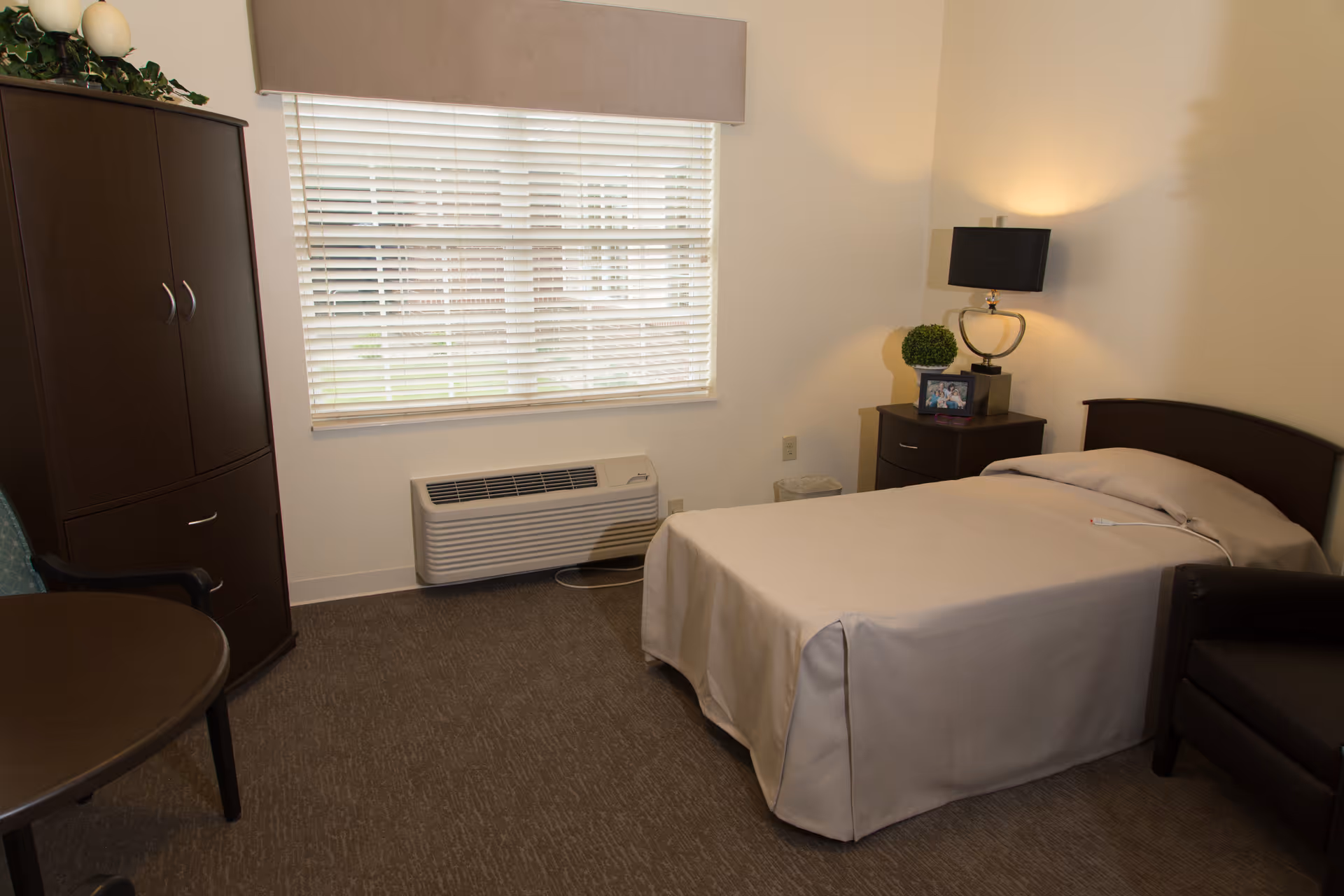 A simple, clean bedroom in a senior living facility featuring a single bed with beige bedding, a dark wooden nightstand with a lamp and a small plant, a dark wooden cabinet, a round table with a chair, and a window with closed blinds letting in natural light.