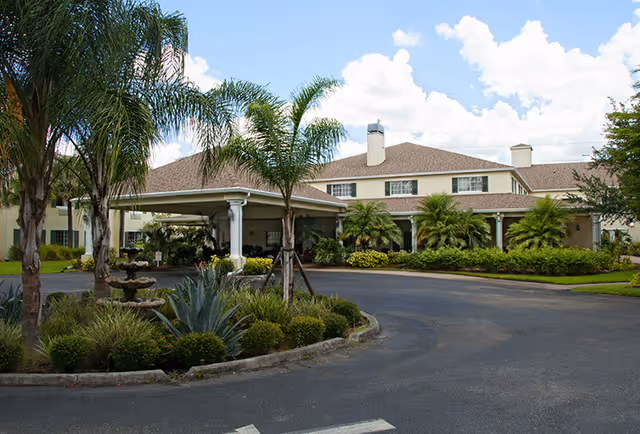 Exterior view of a senior living facility named Lexington Manor featuring a large two-story building with a covered entrance, surrounded by palm trees, shrubs, and a circular driveway under a partly cloudy sky.