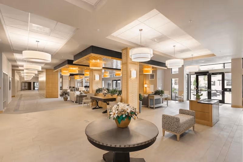 Spacious and well-lit senior living facility lobby with modern decor, featuring a round table with a flower arrangement in the center, comfortable seating areas, wooden columns, large windows, and multiple ceiling light fixtures.