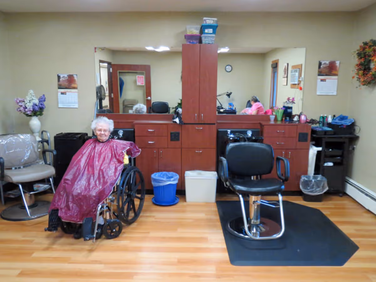 Interior of a hair salon area in a senior living facility with wooden flooring, a large mirror, and cabinetry. An elderly woman in a wheelchair is seated wearing a maroon hairdressing cape. There is a black salon chair on a mat, a vase with flowers, and various salon supplies on the counter.