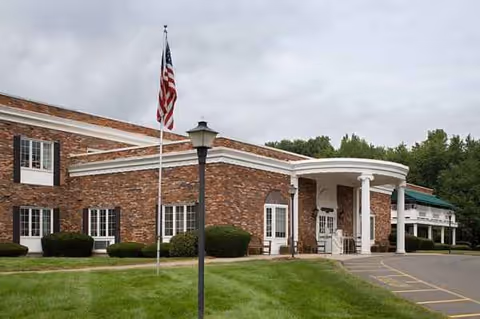 Brick senior living facility with flagpole, entrance pillars, and green lawn.